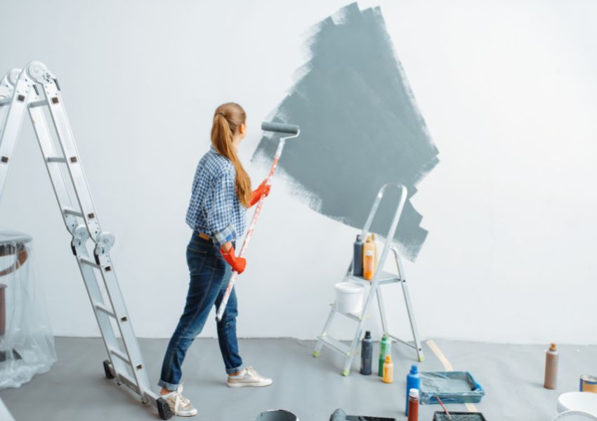 A property manager in jeans and a ponytail paints a rental property wall grey with a roller, as ladders and paint supplies surround them.